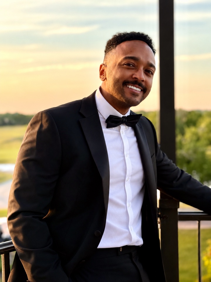 Omar Jean-Baptiste smiling in a black tuxedo in warm sunset light.