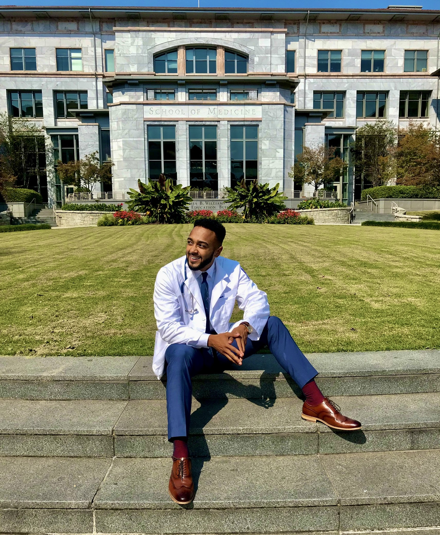 Omar Jean-Baptiste in a white coat, seated on the steps outside a school of medicine.